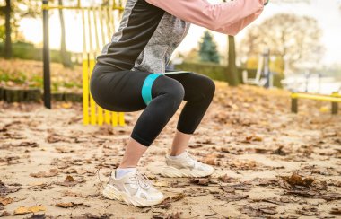 Fitness kızı, sonbaharda basit lastik bantlarla çömelme hareketleri yapıyor. Genç bir kadın parkta egzersiz yapıyor, bacakları yakın.
