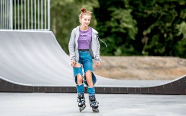 Beautiful girl roller skater riding in city park with ramp. Pretty female teenager rollerskating in casual clothes