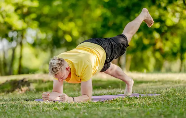Man standing in plank position and practicing yoga outdoors at summer. Guy doing pilates workout for stretching muscles and abs stretght