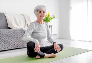 Tatlı Kadın Yoga Mat 'te otururken kameraya bakar, aktif yaşam tarzını somutlaştırır, evde pratik yapar.