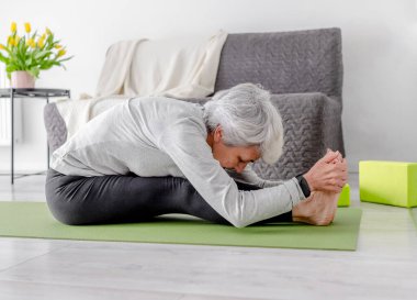 Yaşlı Atletik Kadın Parlak Odada Çiçeklerle Yoga minderi üzerinde otururken Eğiliyor