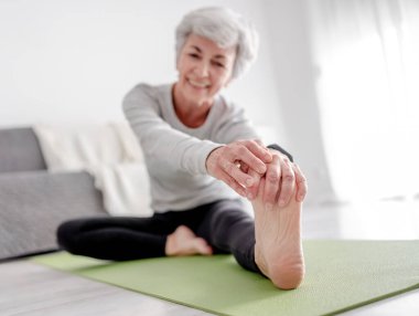 Yaşlı Atletik Kadın Yoga minatında otururken, parlak çiçeklerle dolu bir odada, evinde egzersiz yaparken ayağını uzatıyor.
