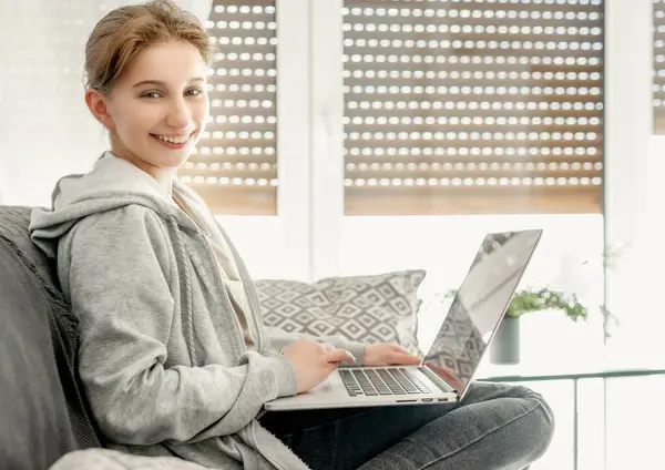 带着笔记本电脑坐在沙发上微笑的女孩 在电视大流行时期用远程教育方案在网上学习 快乐的青少年会花时间在电脑上 — 图库照片#