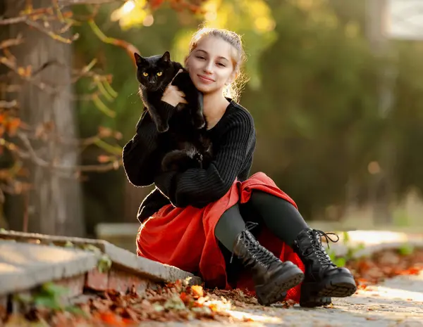 穿着红色裙子的漂亮女孩在外面的街上抱着黑猫 还有秋天的树叶 美丽的少年模特儿与猫科动物坐在公园里 — 图库照片#