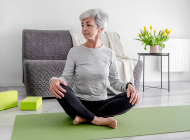 60 yaş üstü bir kadın, parlak bir odada yoga yapıyor.