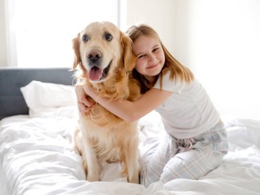 Kız, parlak bir odada sabah yatağında altın av köpeğiyle poz veriyor.