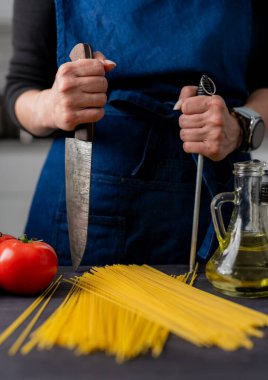 Büyük mutfak bıçağı ve keskinleştirici masanda spagetti, domates, baharat ve fesleğen var.