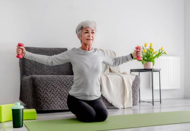 Yaşlı Tatlı Kadın Yoga matında Dambıllarla Egzersiz Yapıyor, Aktif Yaşam Tarzı ve Ev Çalışmalarını Kucaklıyor