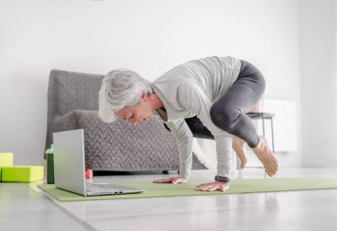 Sevimli 70 yaşındaki kadın laptopla antrenörle kompleks egzersizler yapıyor, evde yoga yapıyor.