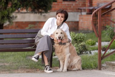 Genç kadın, baharda Golden Retriever 'la evin yanındaki bankta otururken kameraya bakıyor.