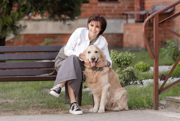 Genç kadın, baharda Golden Retriever 'la evin yanındaki bankta otururken kameraya bakıyor.