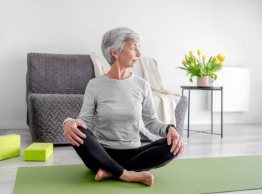 70 yaşındaki kadın Lotus 'ta Yoga yapıyor. Parlak bir odada poz veriyor.