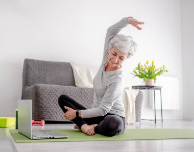 70 yaşındaki hoş kadın antrenörüyle laptopla egzersiz yapıyor, Yoga Mat 'te spor yapıyor.