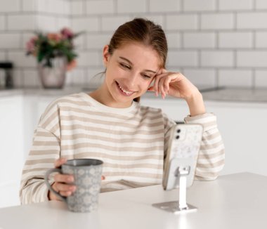 Elinde bir fincan çay olan kız evde akıllı telefondan komik videolar izliyor.