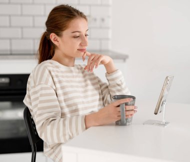 Elinde bir fincan çay olan kız evde akıllı telefondan video izliyor.