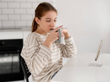 Elinde bir fincan çay olan kız evde akıllı telefondan video izliyor.