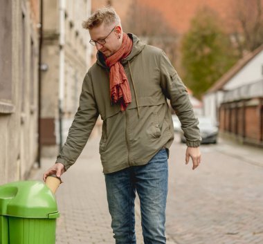 Genç ve şık bir adam şehirde yürüyor ve tek kullanımlık bir kahve fincanını çöpe atıyor.