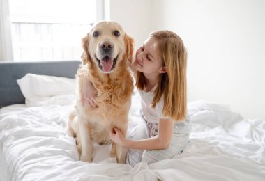 Kız, parlak bir odada sabah yatağında altın av köpeğiyle poz veriyor.