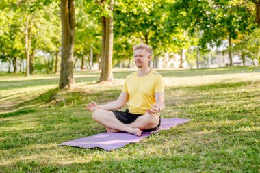 Yazın parkta paspasın üzerinde oturan bir adam yoga yapıyor. Adam kaslarını germek için pilates yapıyor.