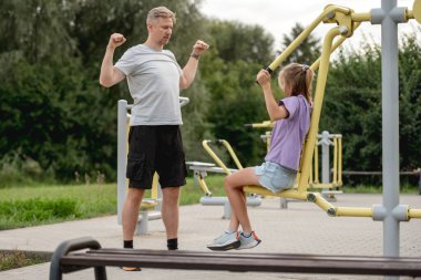 Baba ve kızı oyun alanında birlikte antrenman yapıyor, açık hava aktiviteleriyle kaynaşıyor.