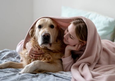 Tatlı küçük kız yatakta bir köpekle oynuyor, battaniyenin altında mutlu bir şekilde sarılıyor.