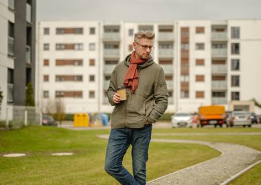 Genç Çekici Adam elinde bir fincan kahveyle bir apartmanın yakınında yürüyor.