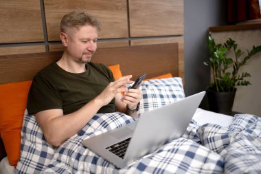 Sabahları, evde dizüstü bilgisayarlı ve akıllı telefonlu bir adam yatakta yatıyor.