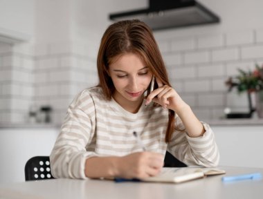 Kız telefonda konuşur ve evdeki deftere yazar.