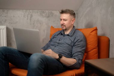 Evde dizüstü bilgisayarla sandalyede oturan yakışıklı adam.