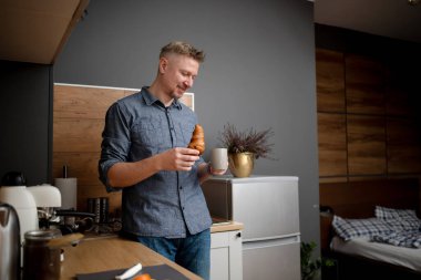 Adam mutfakta kahvaltı için kahve ve kruvasan yiyor.