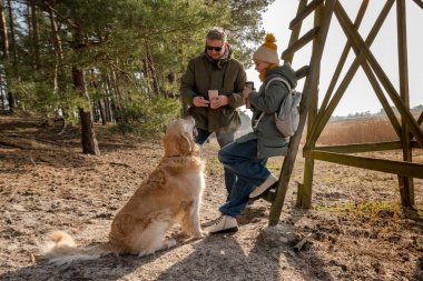 Baba, kız ve Golden Retriever termostan çay içiyorlar.