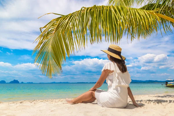 白いドレスと藁帽子を被った若い美しい女性が熱帯のビーチでヤシの葉の下に座っています — ストック写真