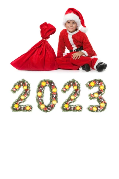 男孩圣诞老人拿着一个圣诞袋和2023年的数字 这些数字是由被白色背景隔开的圣诞树枝做成的 — 图库照片