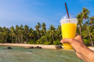 Güneşli bir yaz gününde, tropikal plaj arka planında sarı bir smoothie bardağı tutarken.