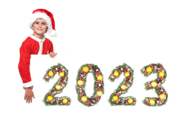 男孩圣诞老人拿着一张圣诞海报和2023年的数字 这些数字是用白色背景隔离开的圣诞树枝制成的 — 图库照片