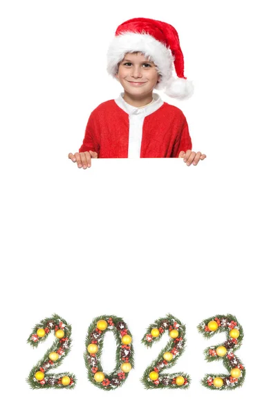 男孩圣诞老人拿着一张圣诞海报和2023年的数字 这些数字是用白色背景隔离开的圣诞树枝制成的 — 图库照片