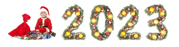 男の子サンタクロースは白い背景に孤立クリスマスツリーの枝によって作られたクリスマスの袋と2023番号を保持 — ストック写真