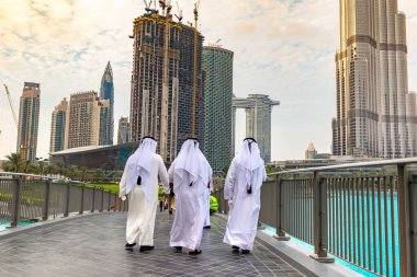 DUBAI, BAE - 5 Nisan 2020: Dubai şehir merkezinde Birleşik Arap Emirlikleri Kulesi 'ne karşı geleneksel beyaz giysili Arap erkekler yürüyor