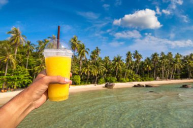Güneşli bir yaz gününde, tropikal plaj arka planında sarı bir smoothie bardağı tutarken.