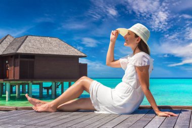 Maldivler adasındaki tropikal sahil iskelesinde (ahşap iskelede) su lüks villalarının önünde oturan güzel genç bir kadın.