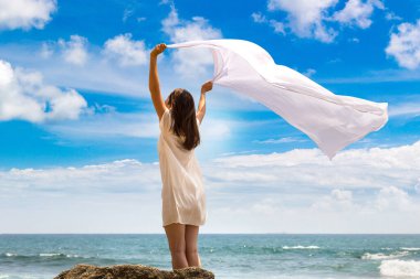Rüzgarda dalgalanan beyaz kumaşlı genç bir kadın portriatı. Denize yakın tropikal bir plajda kalıyor.