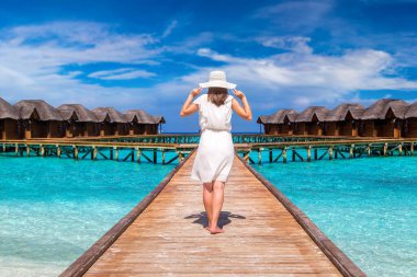 Beautiful young woman in front of water luxury villas standing on the tropical beach jetty (wooden pier) in Maldives island clipart