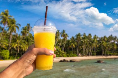 Güneşli bir yaz gününde, tropikal plaj arka planında sarı bir smoothie bardağı tutarken.