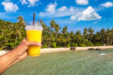 Güneşli bir yaz gününde, tropikal plaj arka planında sarı bir smoothie bardağı tutarken.
