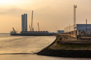 HULL, İngiltere - 18 Şubat 2025: Hull limanı, Siemens Gamesa tesisinde açık deniz rüzgar türbini bileşenleri. Büyük türbin kuleleri, vinçler ve günbatımı ışığı altında yenilenebilir enerjiyi destekleyen bir kargo gemisi.