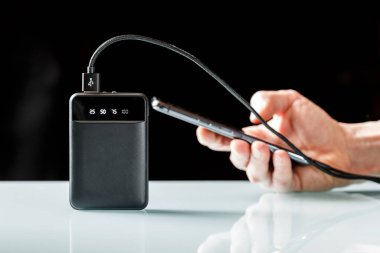 Siyah kablosu olan siyah bir güç bankası bir erkeğin elinde akıllı telefonu şarj ediyor. Ekstra güç kaynağı. Asgari görüntü  