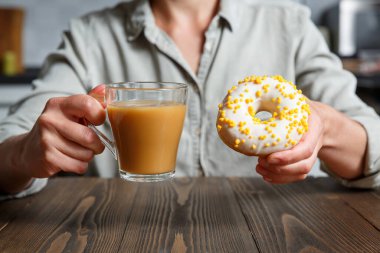 Kadın eli, şeffaf kahve ve donut bardağını beyaz bir jölenin içinde tutuyor ve mutfakta sarı şekerleme serpiştiriyor. Tatlılar, kekler ve çörekler 