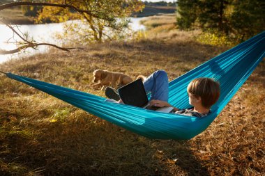 Hammok 'ta dizüstü bilgisayar kullanan genç bir kadın evcil köpeğiyle birlikte gün batımında göle bakıyor. Uzak iş fırsatı kavramı 