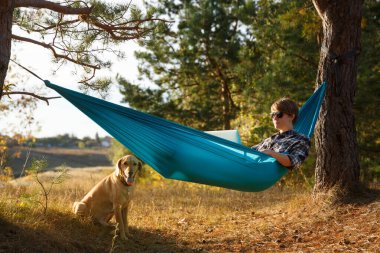 Hammok 'ta dizüstü bilgisayar kullanan genç bir kadın evcil köpeğiyle birlikte gün batımında göle bakıyor. Uzak iş fırsatı kavramı 