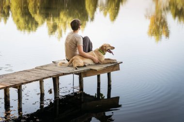 Ahşap bir balıkçı köprüsünde labrador köpeği olan bir kadın. Huzurlu sıcak su manzarasının tadını çıkarıyor. Rahatlatıcı ve evcil hayvanlarla boş zaman.
